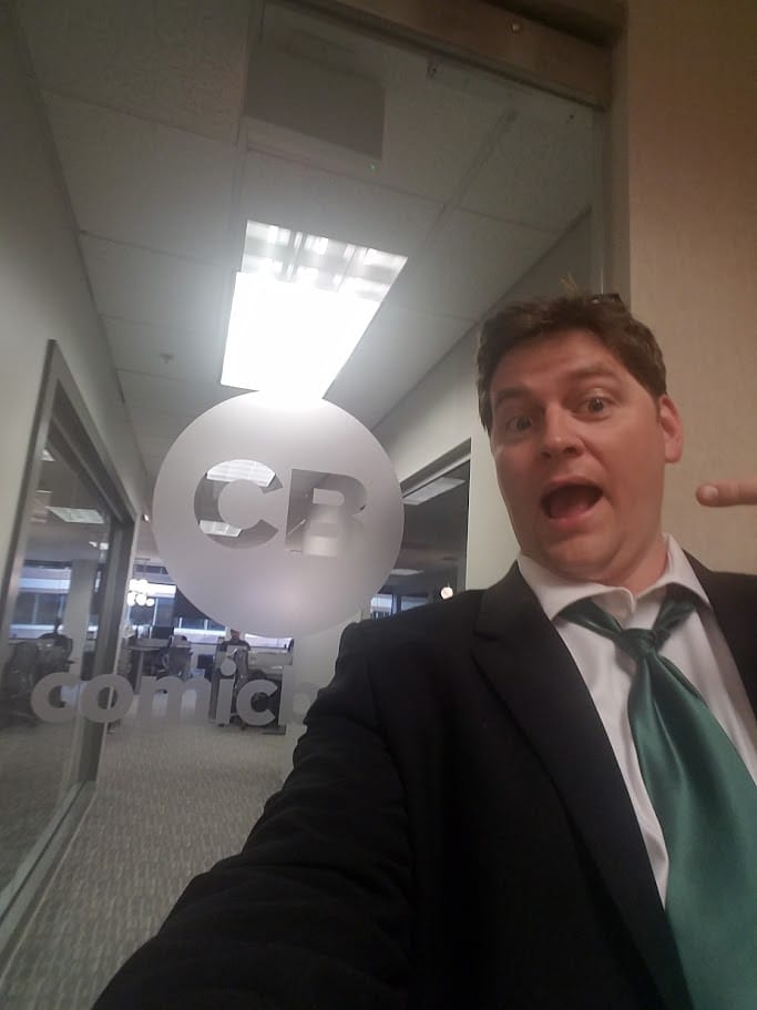Russ in a black suit and green tie, doing a funny face in front of the door to ComicBook.com's office.